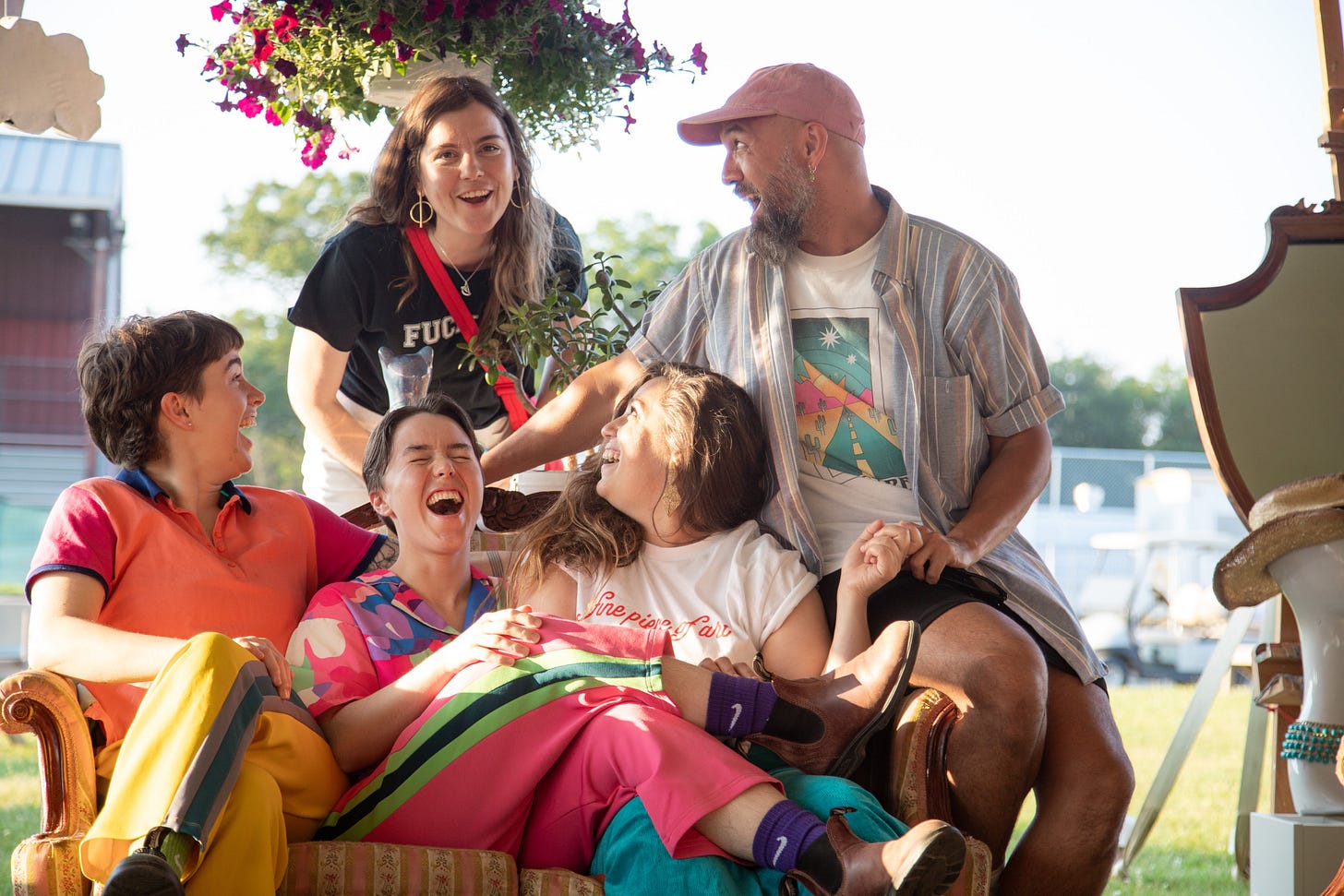 Five people gather around a couch laughing. They are wearing bright colors and gesturing toward each other.