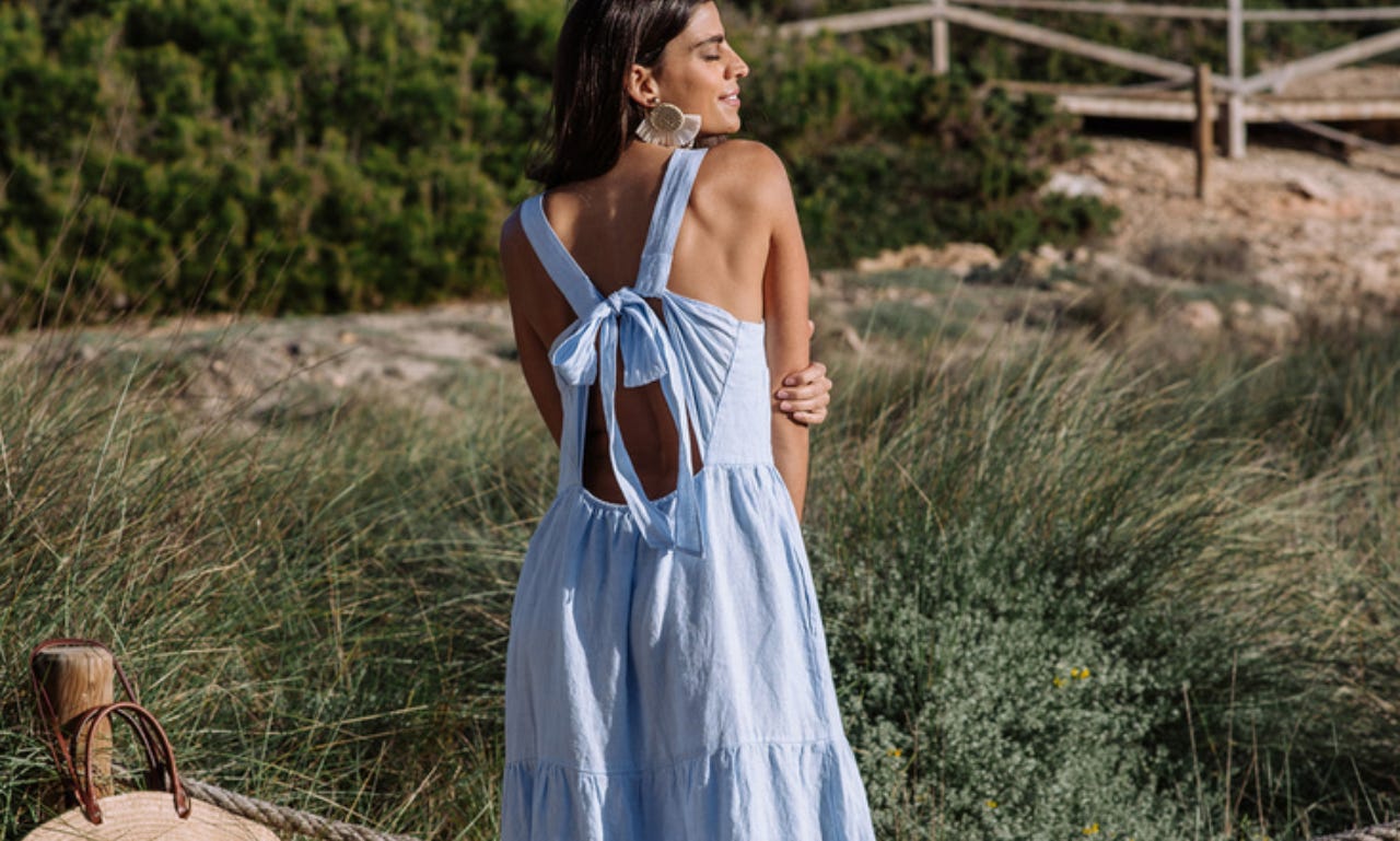 a model stands outdoors besides desert plants and faces away from the camera. she wears a pale blue tiered sundress that is tied between her shoulder blades in a bow, leaving the lower and upper parts of her back exposed.