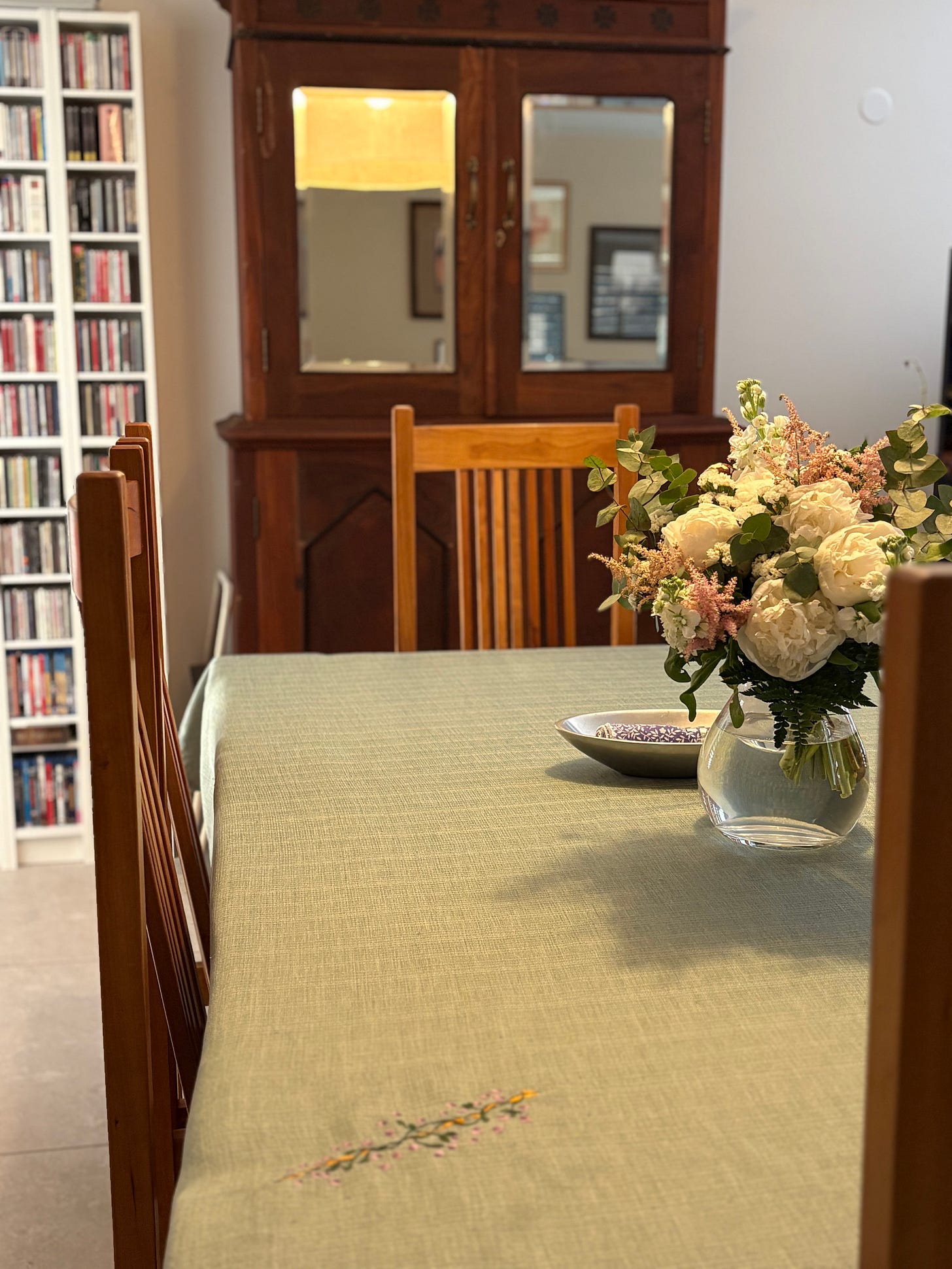 A green tablecloth on a dining table with a vase of peonies, surrounded by Mission-style cherry dining chairs. A teak armoire is in the background.