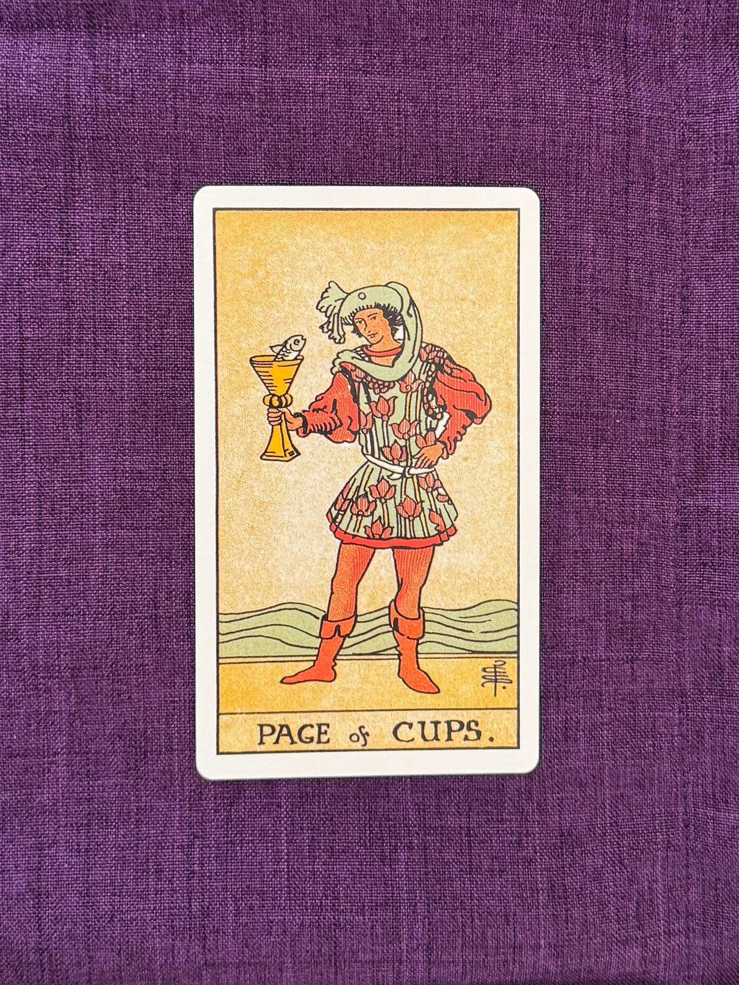 A young figure stands at the water’s edge, holding a golden cup in one hand. A small fish emerges from the cup, its head and upper body visible above the rim. The figure wears a blue tunic decorated with floral patterns, a blue hat, and a long, flowing scarf. The water stretches behind the figure, with low landforms visible in the distance beneath a clear sky.