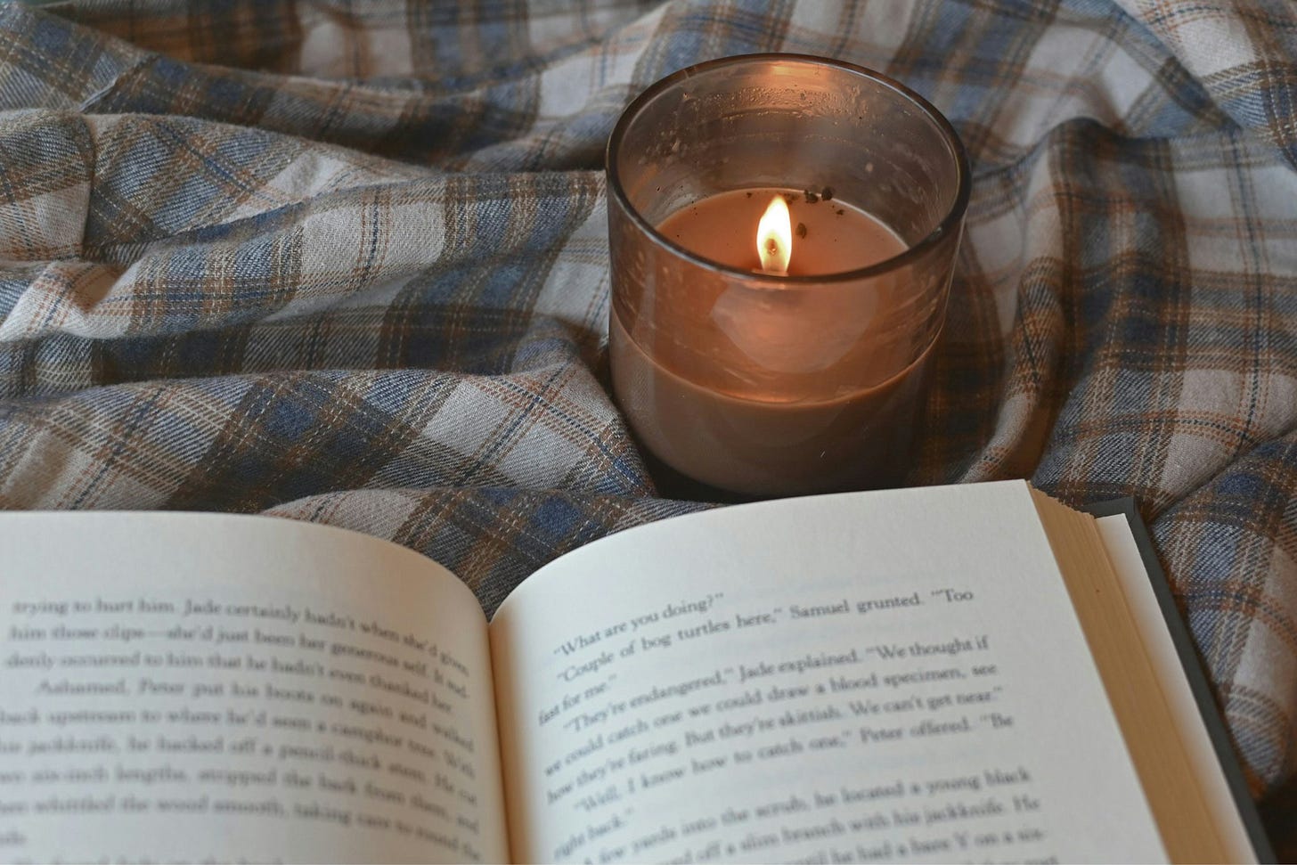 An open hardcover book resting at the bottom of the image, with a lit amber colored candle in a small glass above it, both resting on a rumpled blue, tan, and cream plaid sheet.