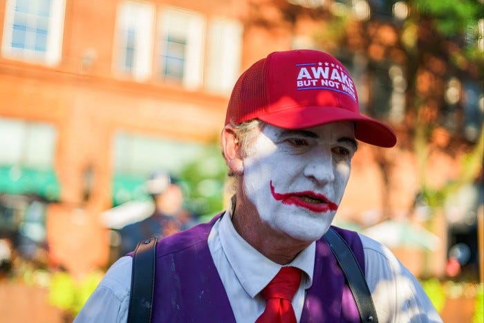 A man with white Joker face makeup and painted red lips wears a red baseball cap with the words Awake but not Woke A man with white Joker face makeup and painted red lips wears a red baseball cap with the words Awake but not Woke