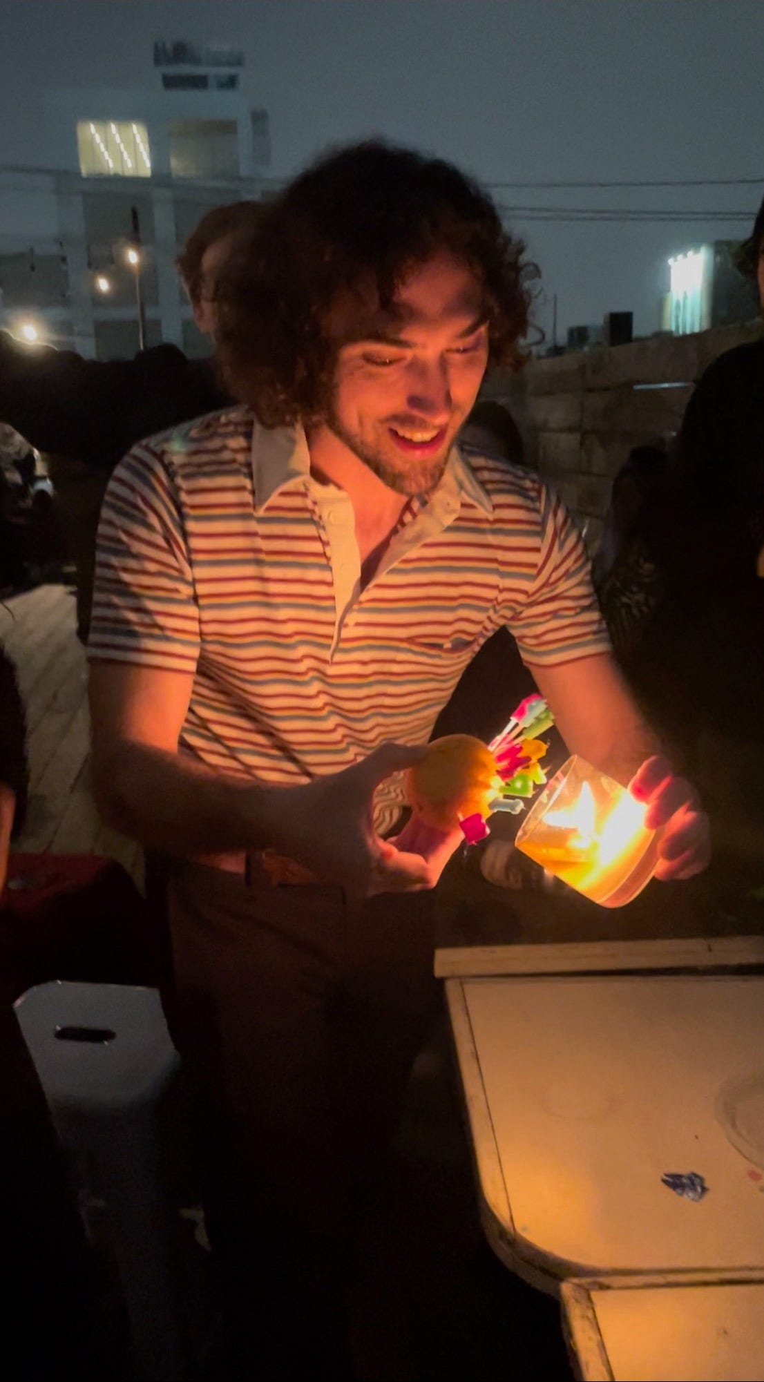 a young white man with a beard lights Happy Birthday candles stuck into an orange on a rooftop at night