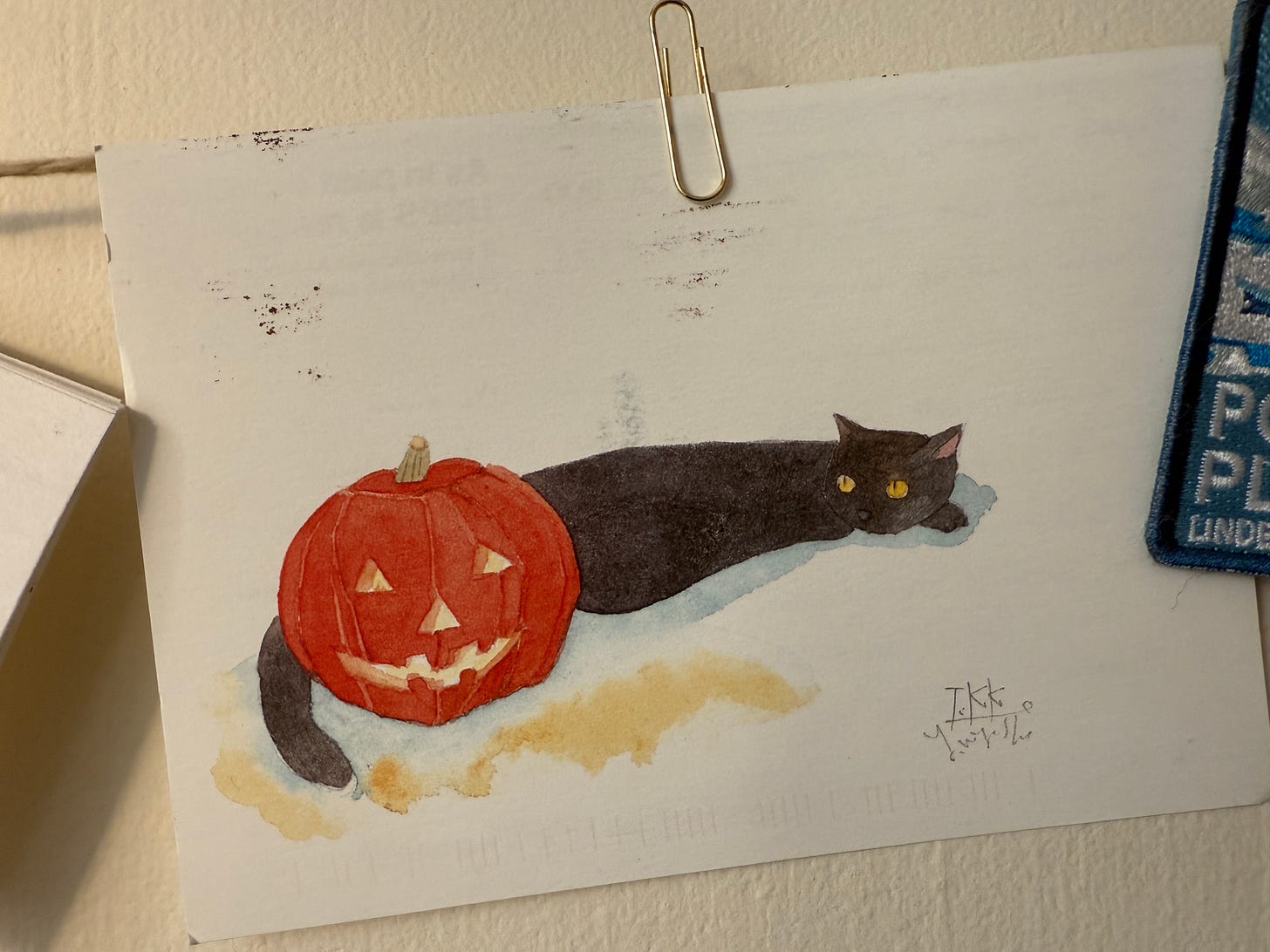a close up of a postcard hanging on a string on a wall. The postcard features a black cat laying beside a pumpkin that's been carved into a classic Jack-o-lantern.