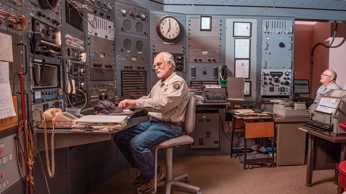 photo of two uniformed men sitting in control center with huge analog machinery bank in background