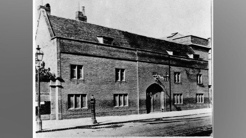 A black and white photograph of a now-demolished building known as the Spinning House in Cambridge. It is brick built with mullioned windows on its ground and first storeys. There are small dormer buildings in its pitched roof. It has a large arched doorway with a door made of wooden planks.