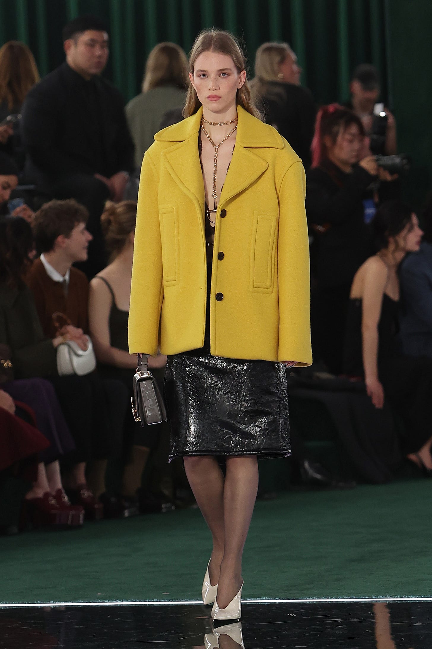 A blonde model walks down a runway wearing an oversized yellow cloth coat and a black patent leather skirt.
