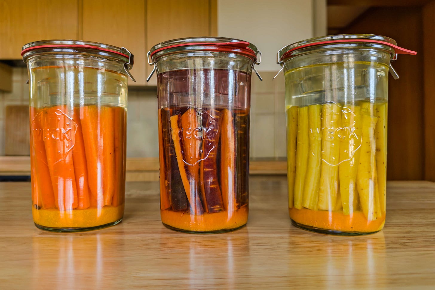 Drei verschlossene WECK-Gläser mit fermentierten Karotten-Sticks in verschiedenen Farben – orange Karotten, violette Purple Haze und gelbe Pfälzer Karotten – stehen auf einem Holztisch in einer Küche.