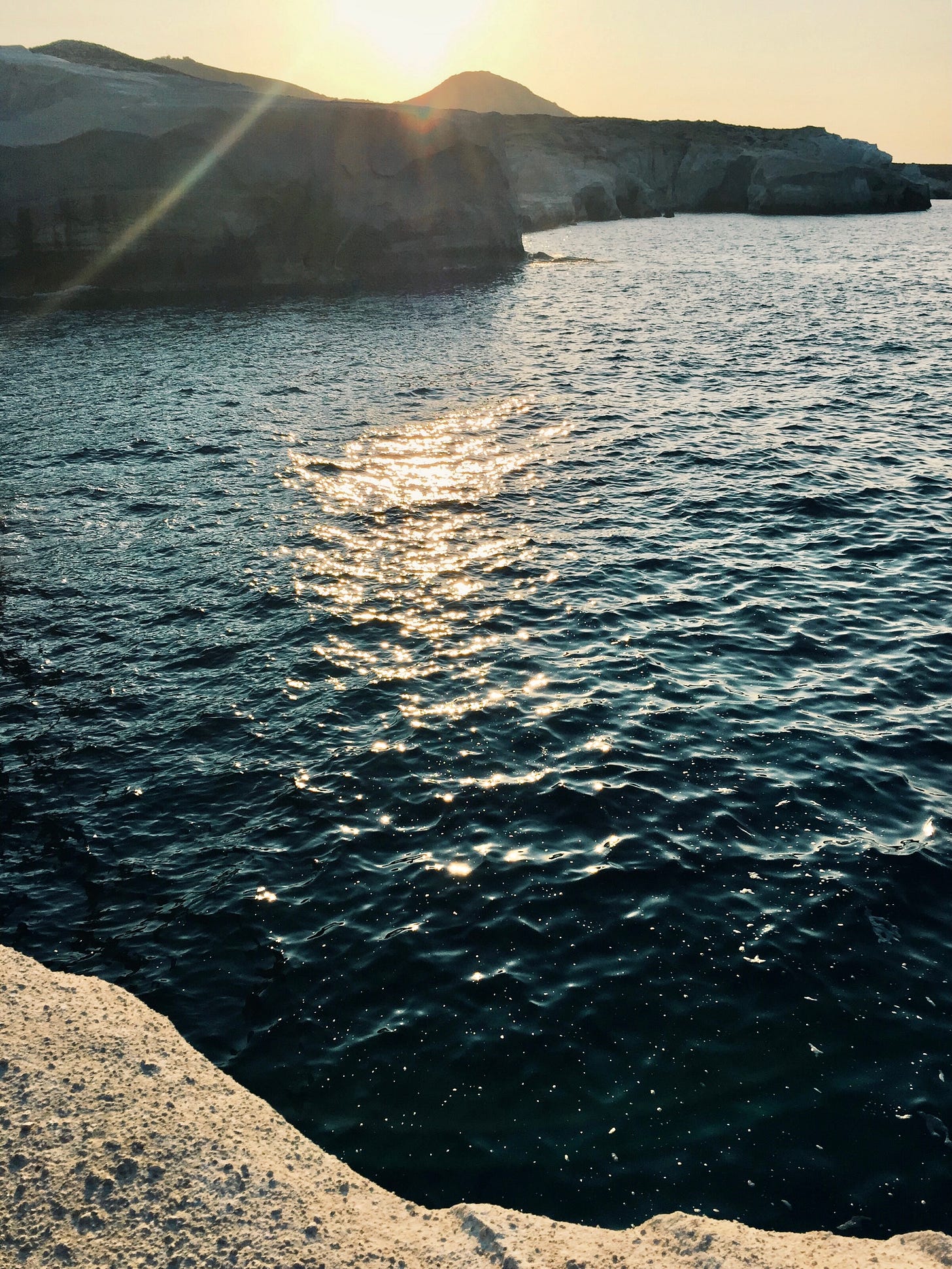 The sun sets behind white cliffs and a deep blue ocean on the island of Milos, Greece casting a warm glow
