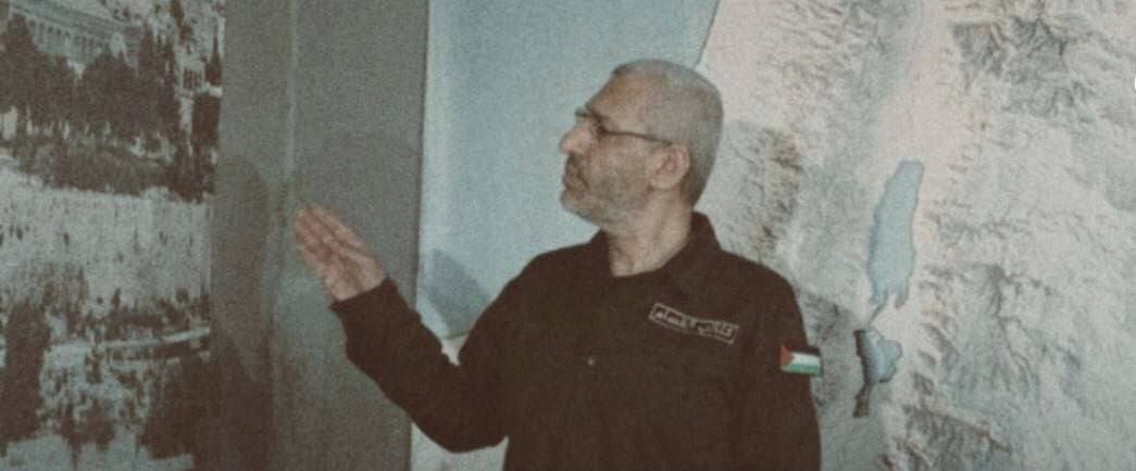 Elderly man with gray hair beard and glasses wearing black jacket with Palestinian flag patch on sleeve stands in front of light gray wall with faded black-and-white mural depicting indistinct figures gesturing with right hand extended palm open image has vintage faded color tone