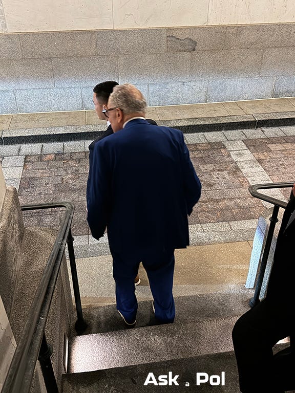 A politiican in a blue suit and a security officer in a black suit await a car in what looks like a tunnnel at US Capitol 
