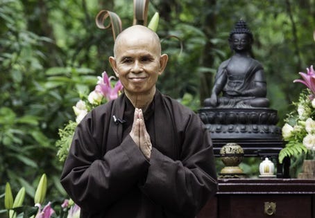 Thich Nhat Hahn standing with hands in prayer position in front of trees and flowers and a Buddha statue Thich Nhat Hahn standing with hands in prayer position in front of trees and flowers and a Buddha statue