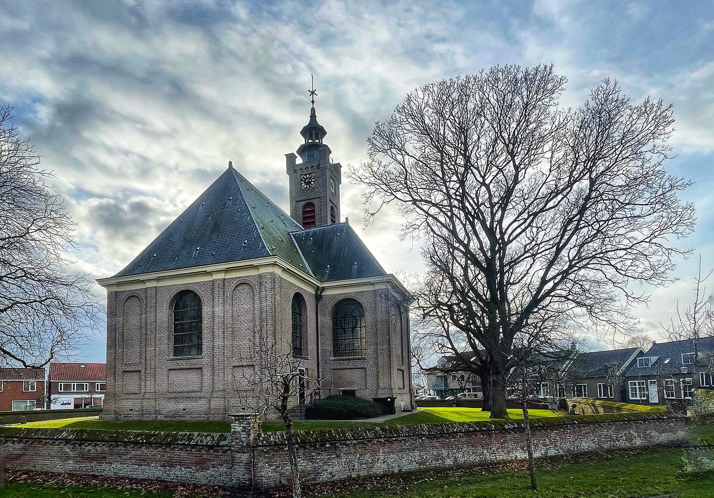 A small church and a huge tree next to it. It’s in the center of a village, on the left and right you can see houses around the church square.