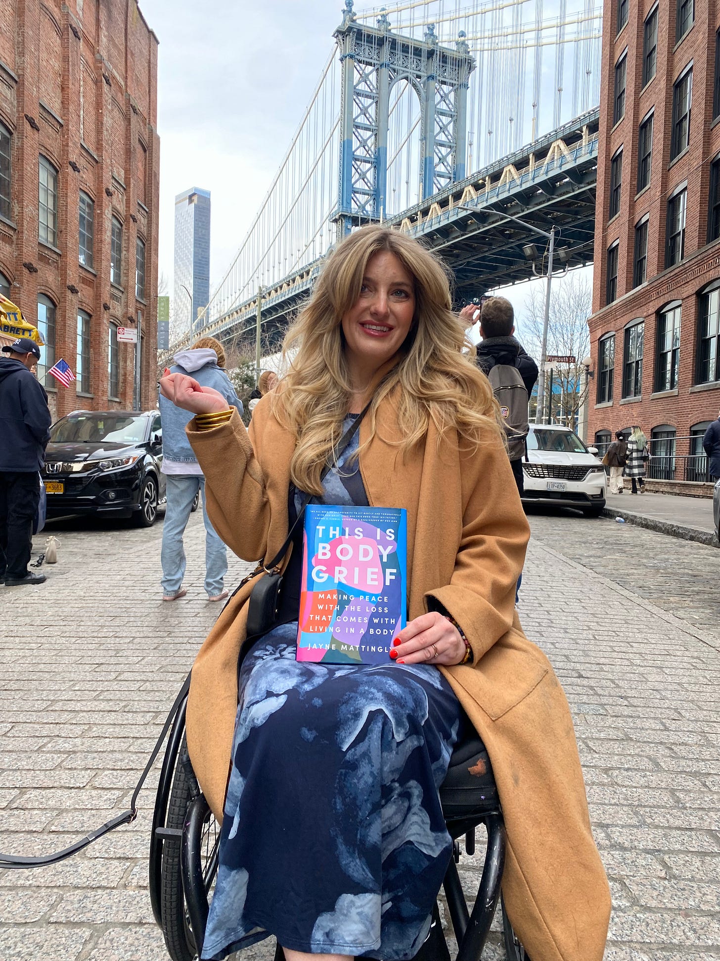 Jayne in her Wheelchair in front of the bridge in Dumbo posing with her book THIS IS BODY GRIEF.