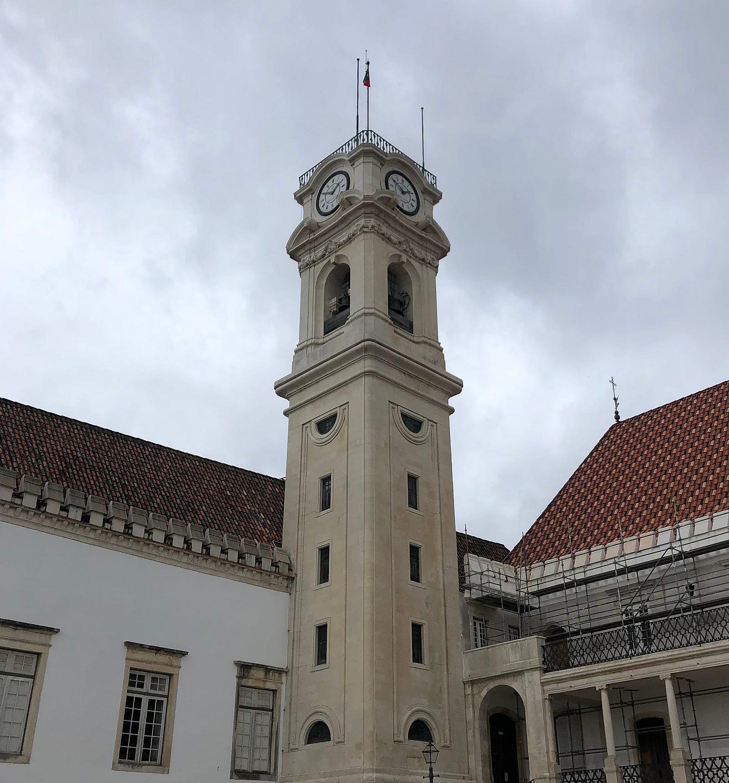 A photo of the cream-coloured stone clock tower at The University of Coimbra in Portugal, adjoined by white plastered buildings with terracotta-tiled roofs, set perpendicularly, all set against a grey, cloudy sky. A photo of the cream-coloured stone clock tower at The University of Coimbra in Portugal, adjoined by white plastered buildings with terracotta-tiled roofs, set perpendicularly, all set against a grey, cloudy sky.