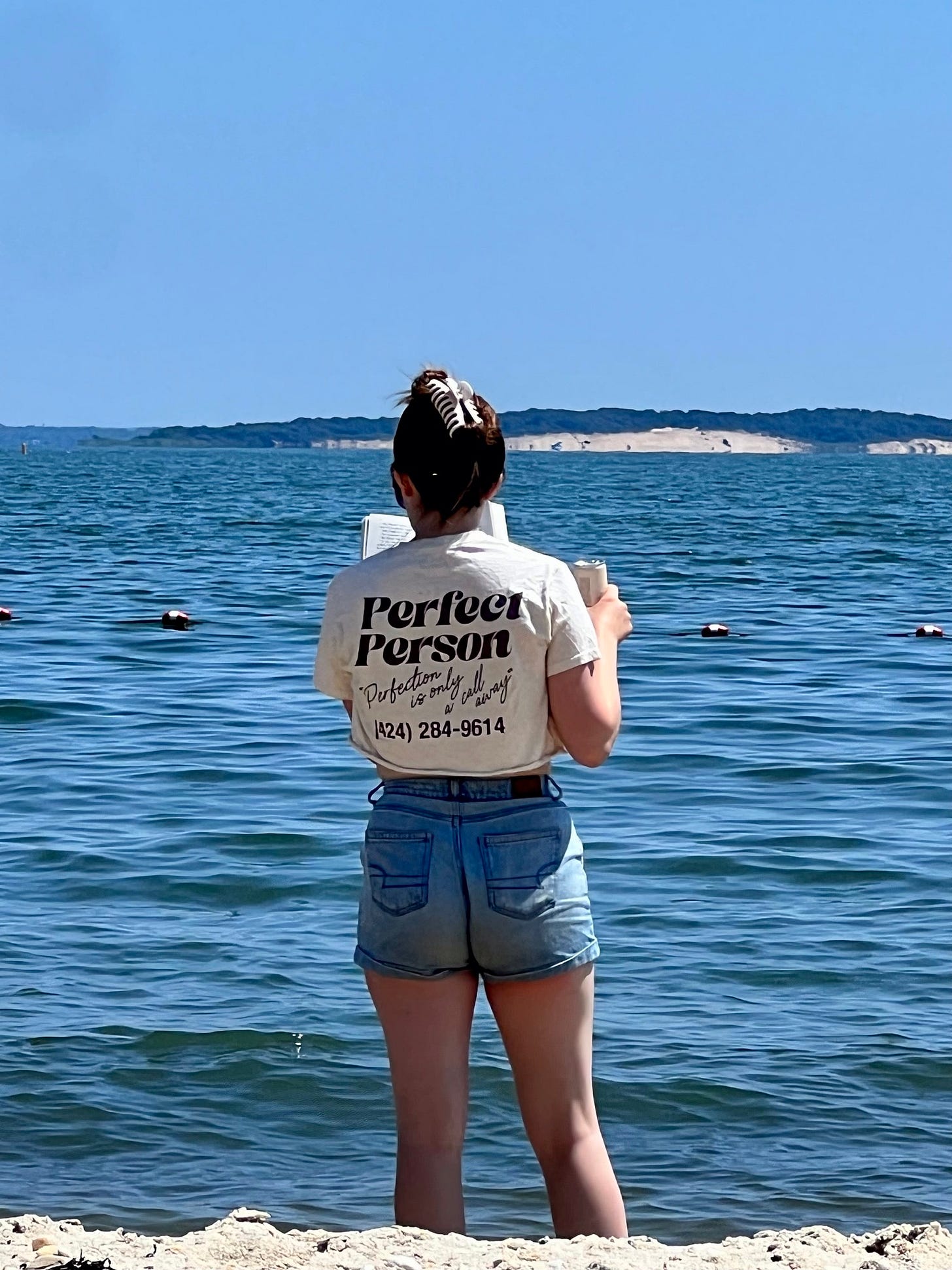 A young woman on a beach standing in front of the water with her back to the camera, with a book in one hand and a drink in the other. Her shirt reads: PERFECT PERSON/ "Perfection is only a call away" / (424) 284-9614)