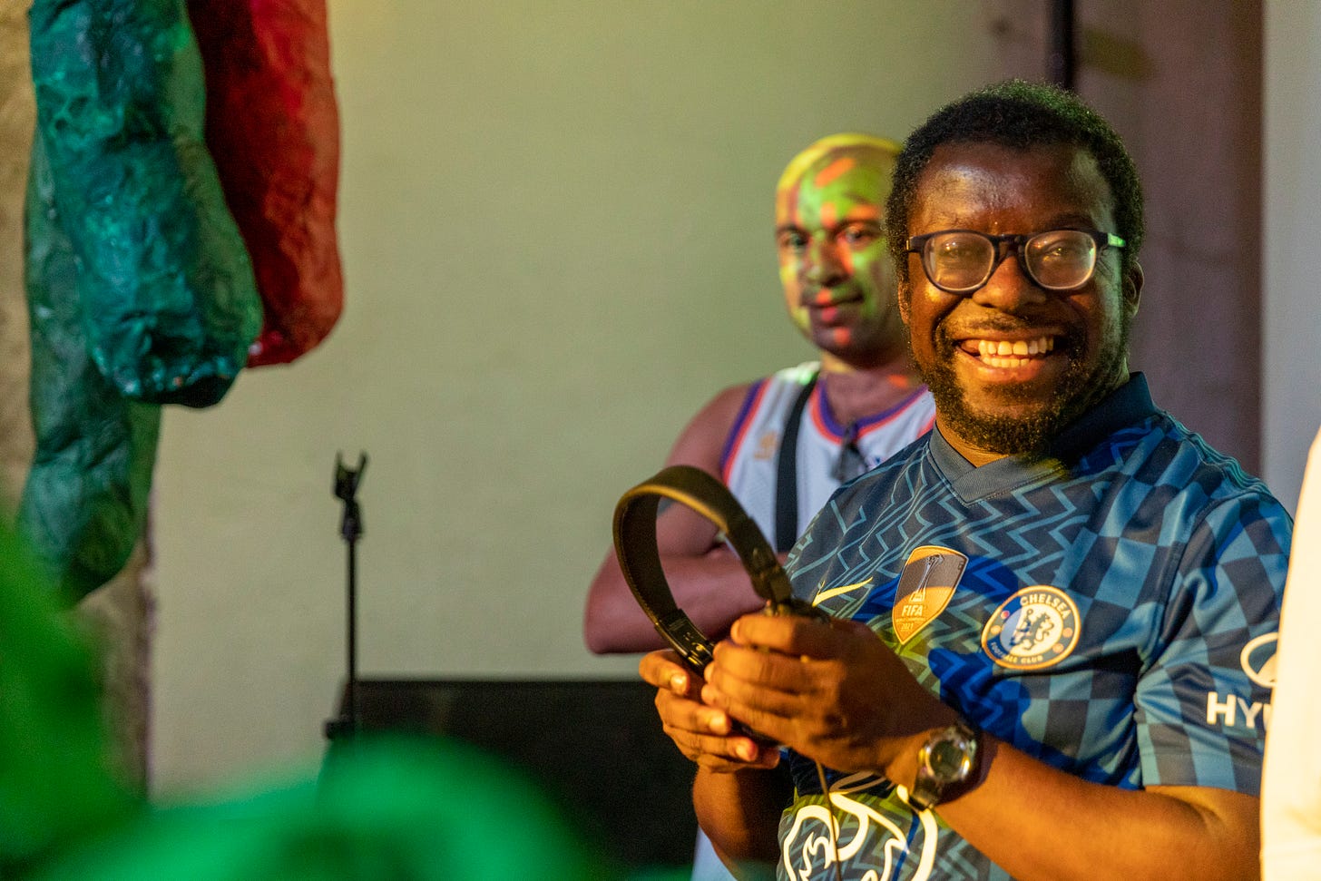 A Black man is smiling at the camera, holding headphones in a well-lit club.