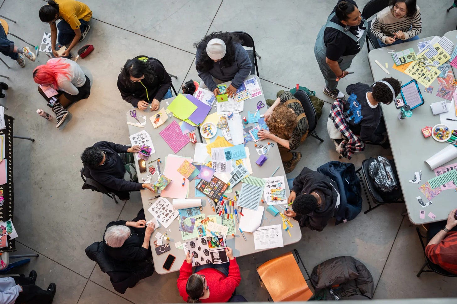 Overhead view of people gathered around tables making zines with paper, scissors, and colorful materials. Overhead view of people gathered around tables making zines with paper, scissors, and colorful materials.