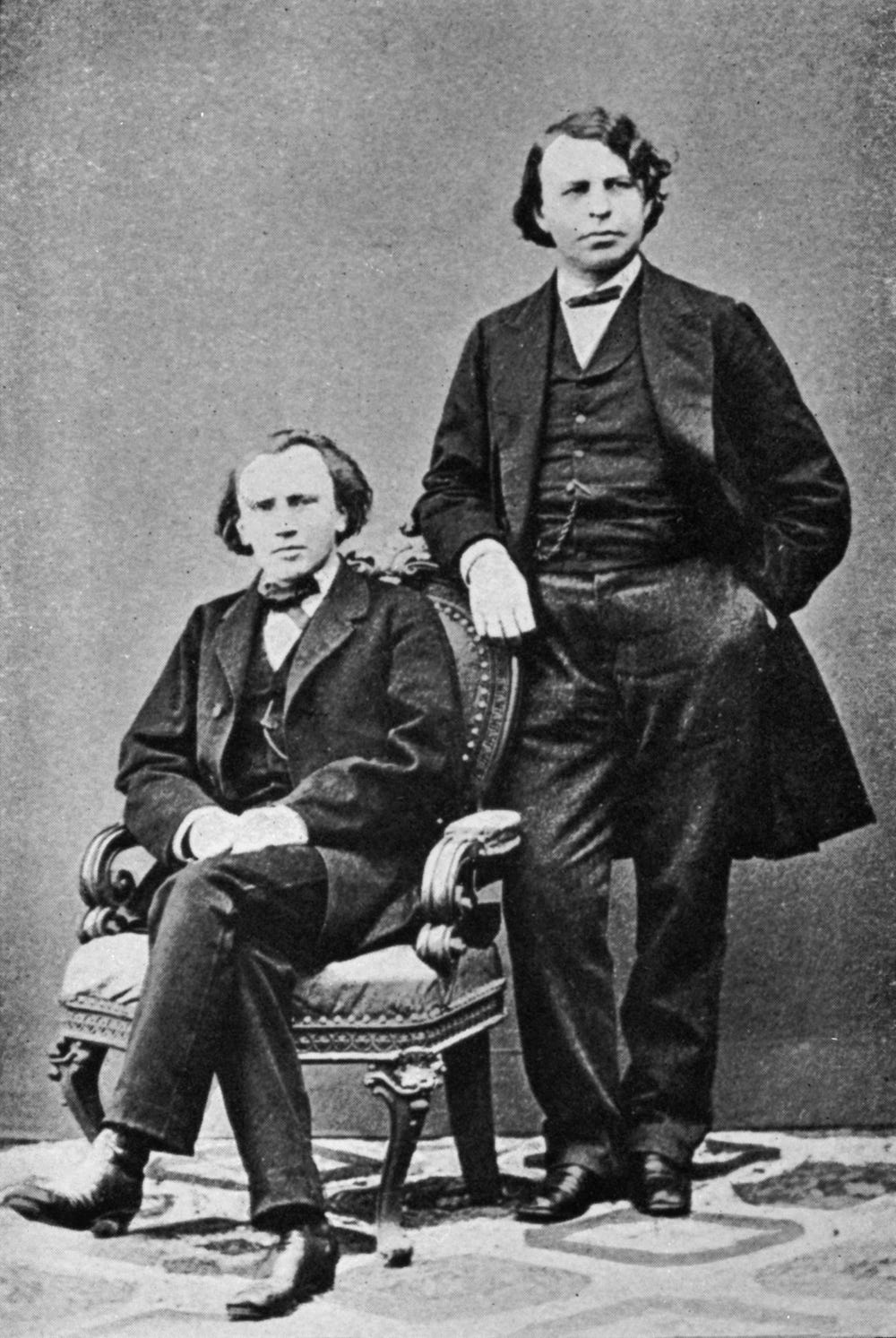 Black and white photo from the 19th century of two men in matching suits, one seated in a chair and the other standing beside him