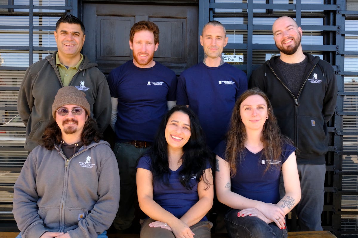 seven staff members pose in two rows; a shopfront can be seen in the background
