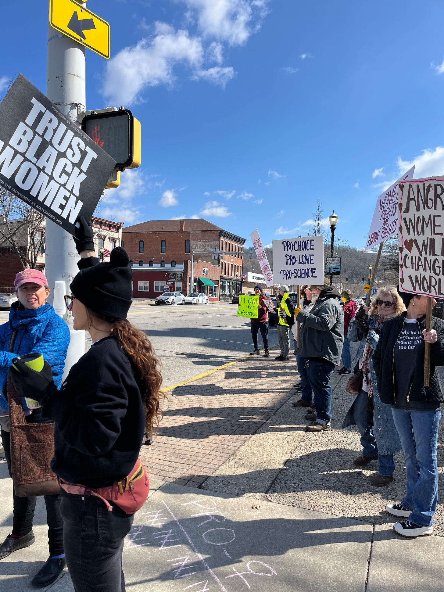 A group of people lined up along a small town street, holding signs that say things like Trust Black women and Pro-choice, pro-love, pro-science
