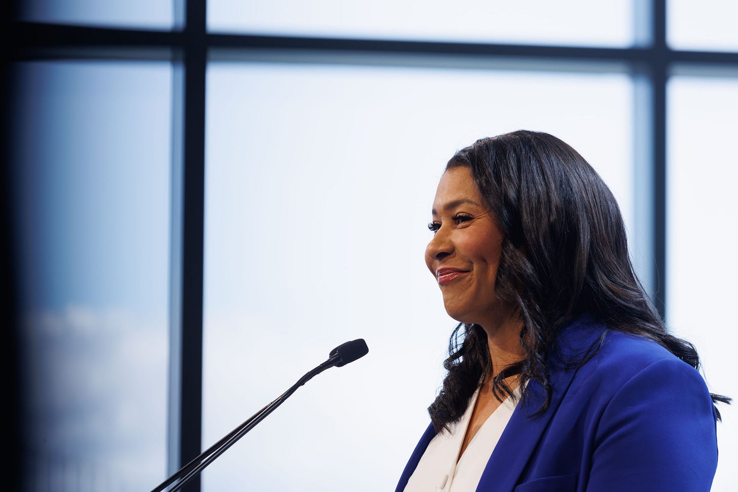 A smiling woman in a blue blazer stands at a podium with a microphone, in front of a window with a blue sky.
