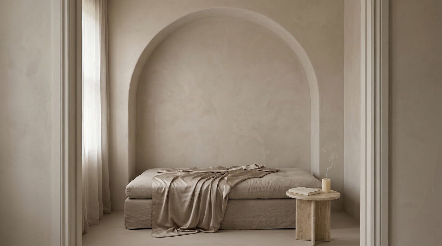 A linen daybed with a rumpled matte champagne satin throw pulled halfway back inside a plaster arch alcove, with a small travertine side table holding a closed cream journal and a candle with a thin wisp of smoke rising, diffused light filtering through sheer curtains.
