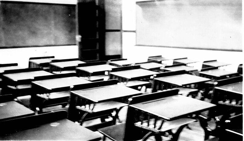 Black and white photograph of an empty classroom full of wooden desks