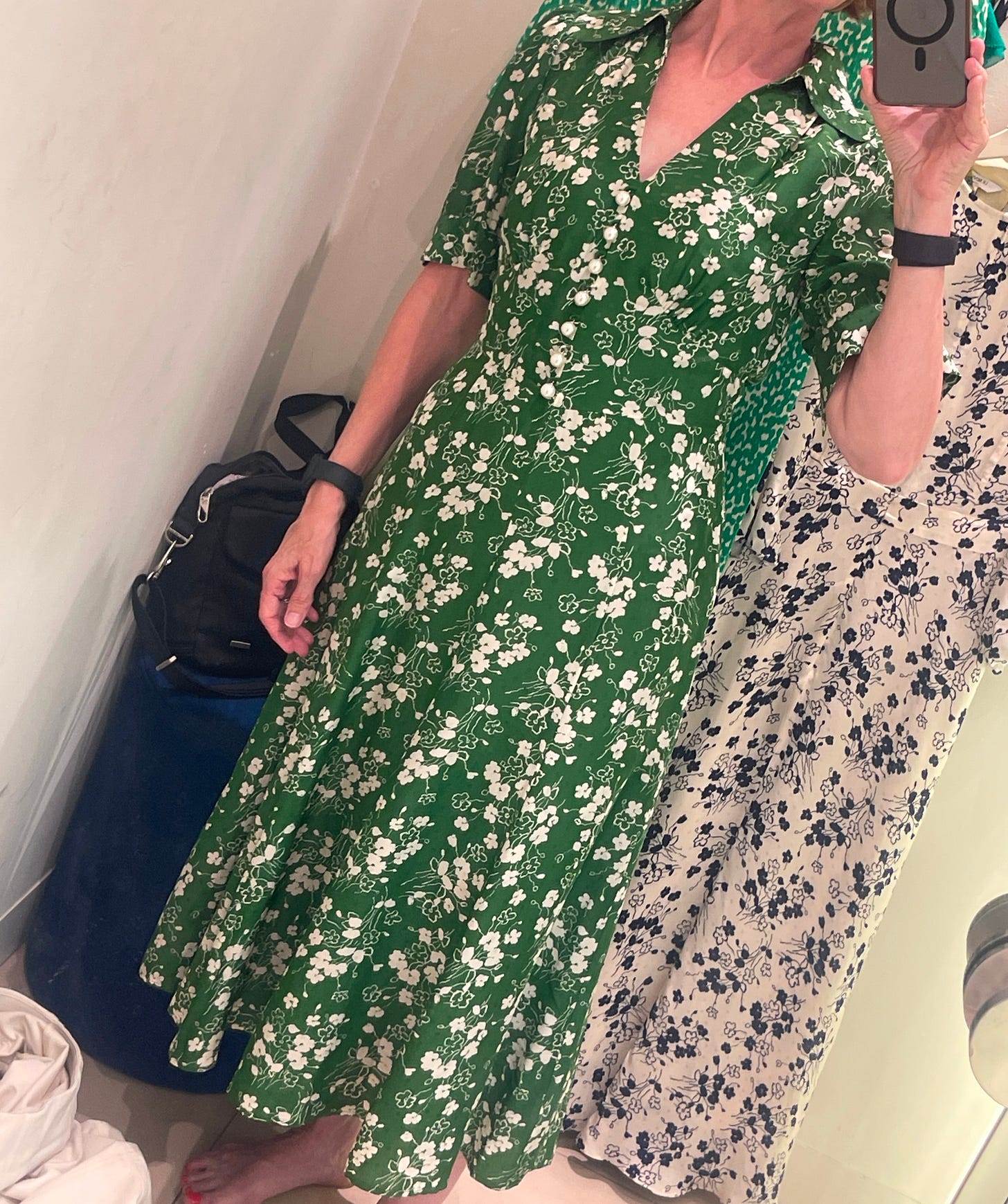A woman stands barefoot in a fitting room wearing a green floral LK Bennett dress with pearl buttons, taking a mirror selfie. Other dresses hang in the background.