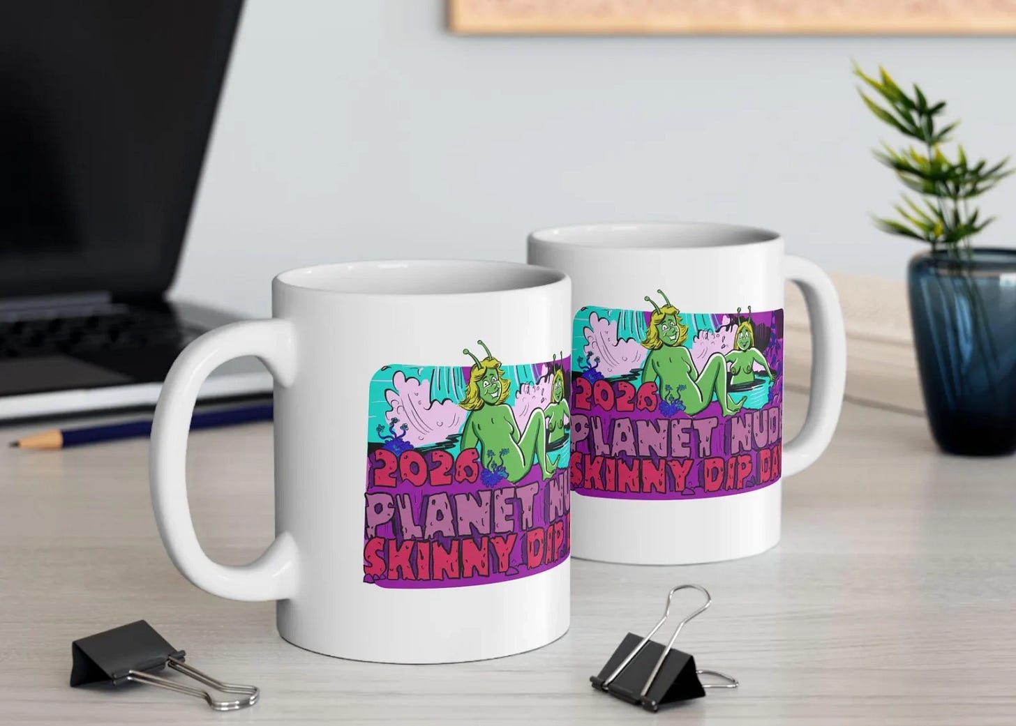Two white ceramic mugs on a wooden desk, both printed with the 2026 Planet Nude Skinny Dip Day art, two green antennaed figures relaxing in a turquoise spring. A laptop, a pencil, binder clips, and a small plant sit nearby. Two white ceramic mugs on a wooden desk, both printed with the 2026 Planet Nude Skinny Dip Day art, two green antennaed figures relaxing in a turquoise spring. A laptop, a pencil, binder clips, and a small plant sit nearby.