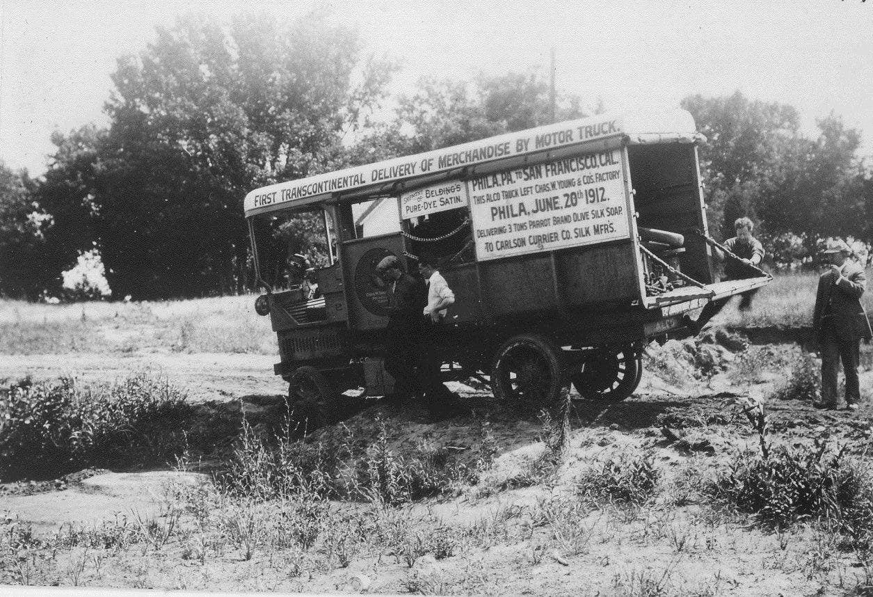 Picture of the first transcontinental motor truck shipment in the US. An old motor truck is stuck in a dirt road, and four men are working to take it off Picture of the first transcontinental motor truck shipment in the US. An old motor truck is stuck in a dirt road, and four men are working to take it off