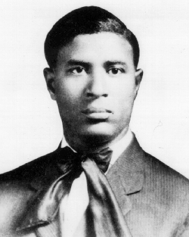 Black-and-white portrait photograph of a young Garrett Morgan, head and shoulders, wearing a dark suit jacket with a wide, light-colored ascot tie knotted at the throat. He has short dark hair combed close to his head and a neutral expression, facing the camera directly.