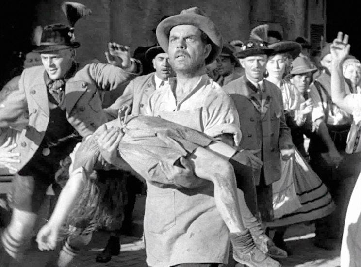Maria's father arrives in town, carrying the body of his drowned daughter, his face a mask of grief. Behind him townspeople, in peasant dress gather. A still from 1931 film Frankenstein