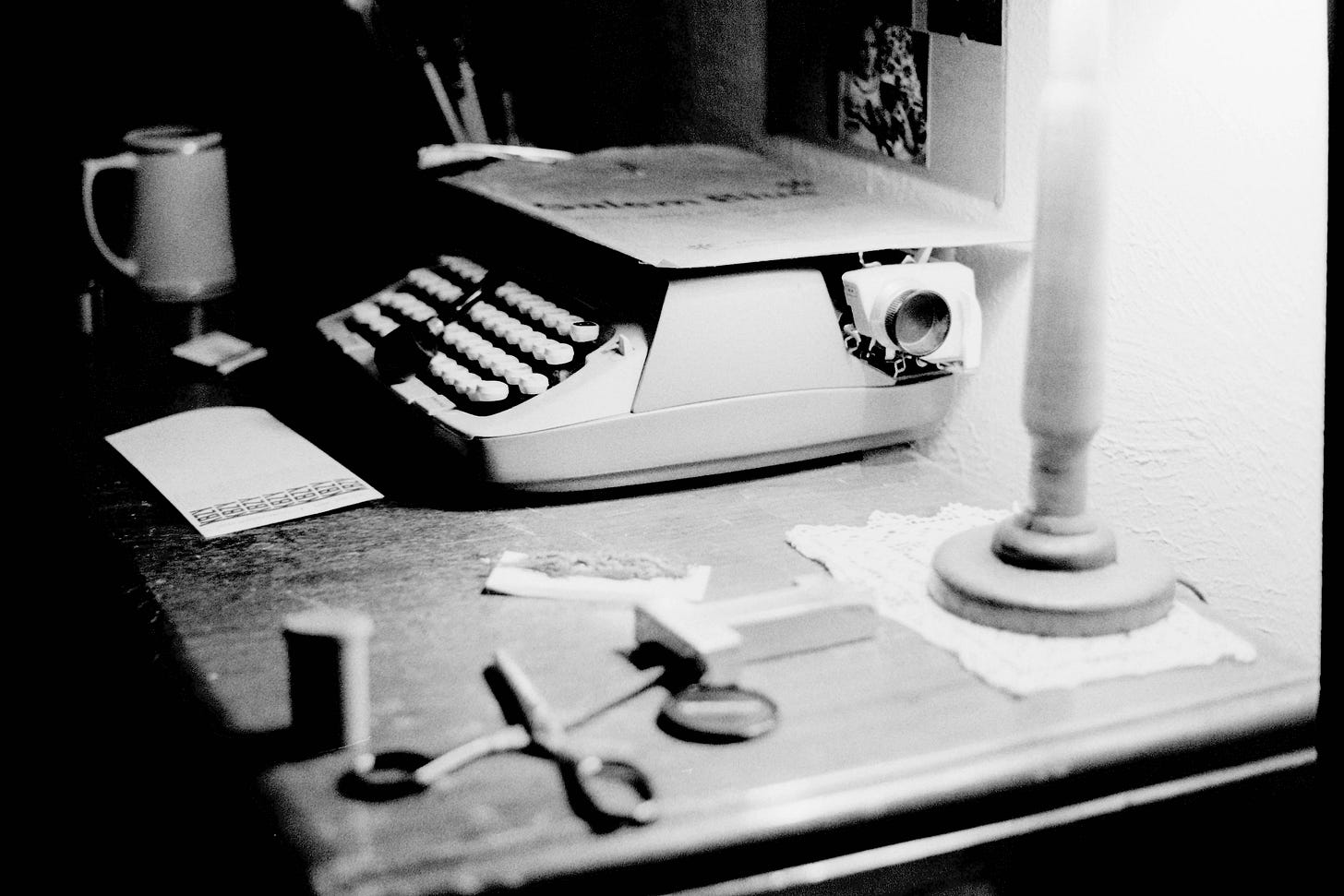 A black-and-white photograph of a vintage writing desk illuminated by a small lamp. At the center sits an old typewriter with a sheet of paper loaded, surrounded by scattered objects including scissors, a roll of film, a coffee mug, and a notepad. The soft light and shallow depth of field create a nostalgic atmosphere, evoking the quiet focus of a writer’s workspace from decades past.