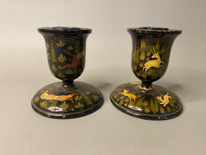 May include: Two antique candle holders with a black base and a flared top. The candle holders are decorated with colorful painted scenes of animals and foliage. One features a running deer and the other a running rabbit. The design is intricate and detailed.
