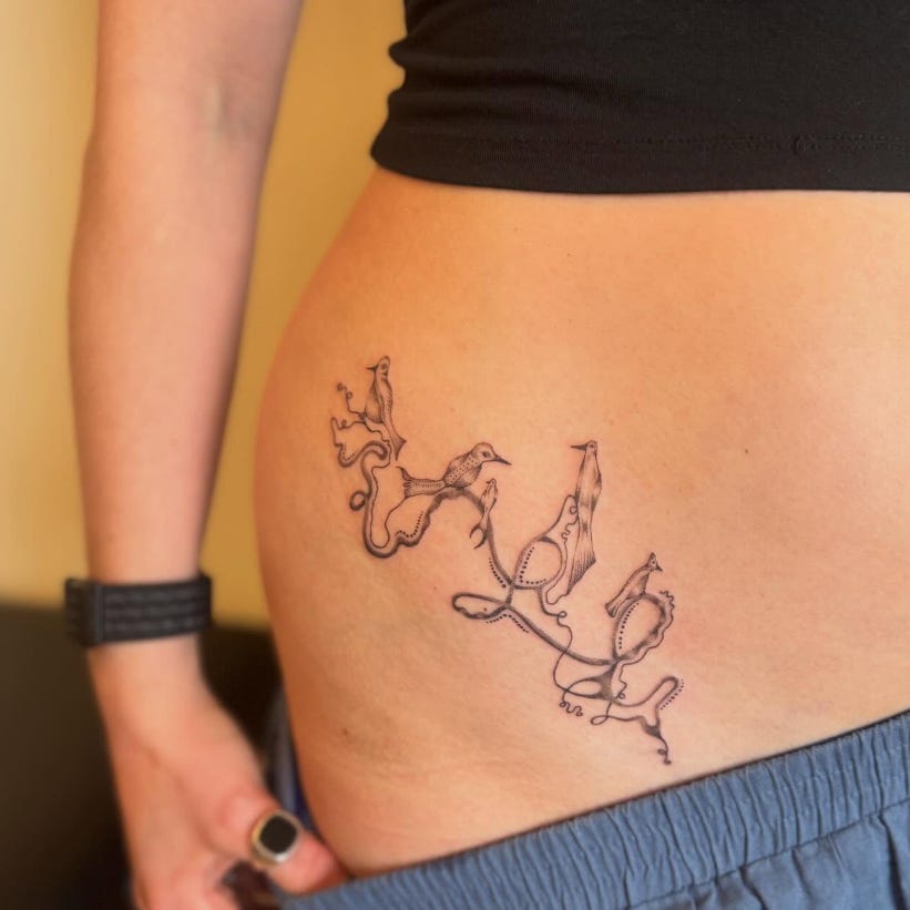 Hand holding pants down to reveal the hip and the area above it. A tattoo in black ink of birds perched atop branch-like flowing lines curves over the hip bone.