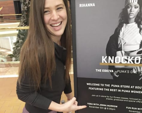 Three side-by-side images of a woman leading movement and healing-centered workshops. Left: she smiles while standing next to a Puma x Rihanna event sign, indicating a yoga class collaboration. Center: she poses with a diverse group of students proudly holding up certificates and drawings from a workshop, surrounded by papers on the floor. Right: she kneels on a yoga mat, gently holding a student’s hands in a moment of somatic connection and support.