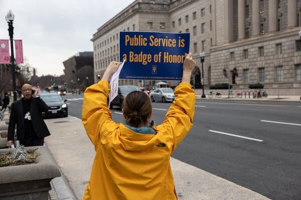 A woman, shown from behind, wearing a yellow jacket and holding a sign reading “Public Service is a Badge of Honor!" over her head. A woman, shown from behind, wearing a yellow jacket and holding a sign reading “Public Service is a Badge of Honor!" over her head.