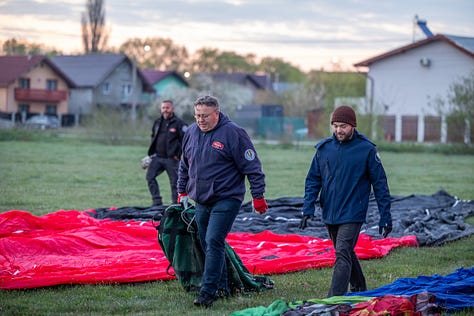Înainte ca baloanele să urce, totul începe jos, pe iarbă, în frigul subțire al dimineții sau în lumina care abia se stinge peste câmp. Oamenii își ocupă locurile, verifică arzătoarele, întind pânza, ridică nacela, privesc cerul și, mai ales, ascultă vântul. În jur, mașinile stau cu farurile aprinse.