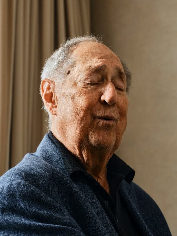 An older man with gray hair in a blue jacket over a black shirt closes his eyes in contemplation.