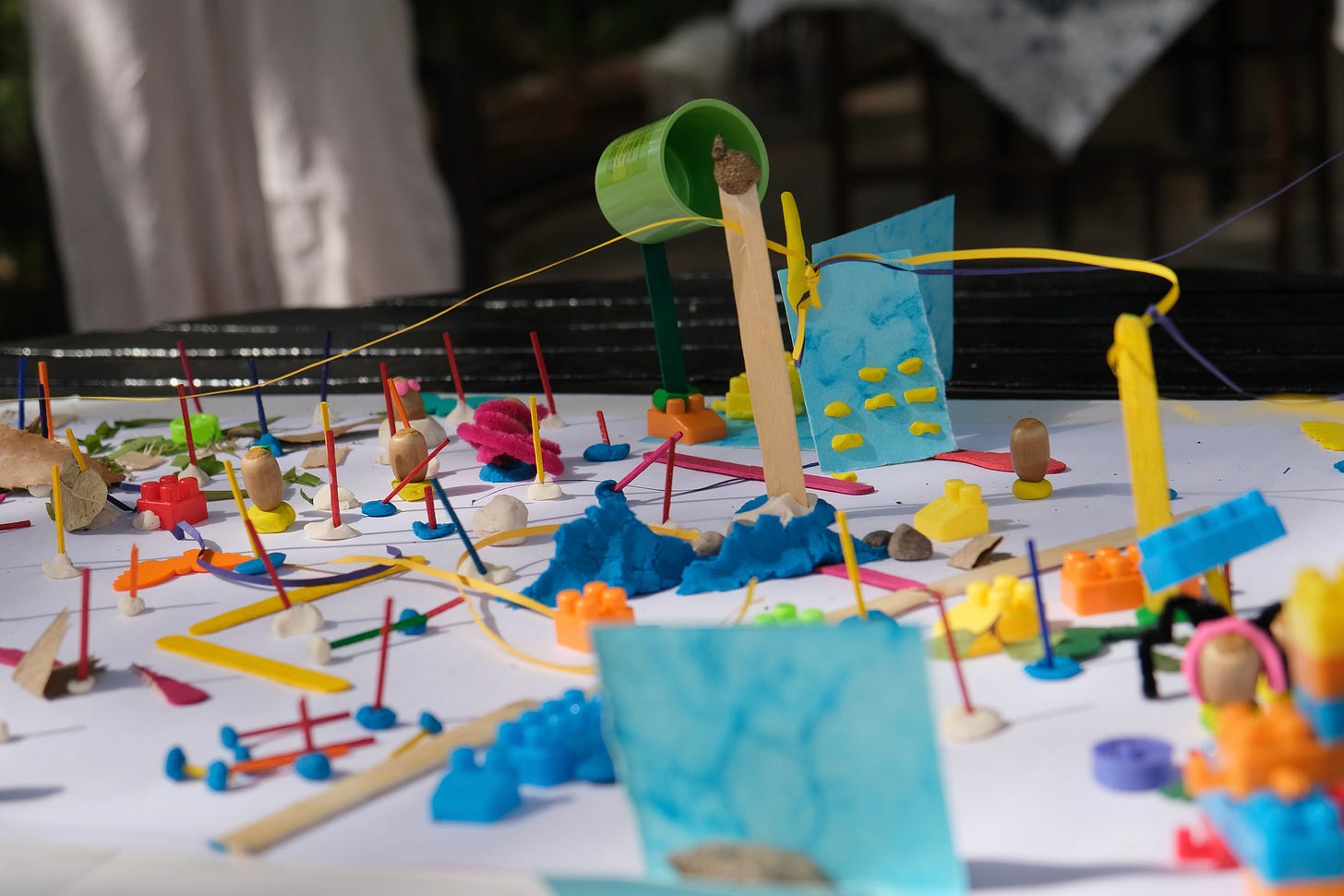A white sheet of paper with a variety of small trinkets like lego pieces, playing dough, leaves, ice cream sticks, quilling paper etc.