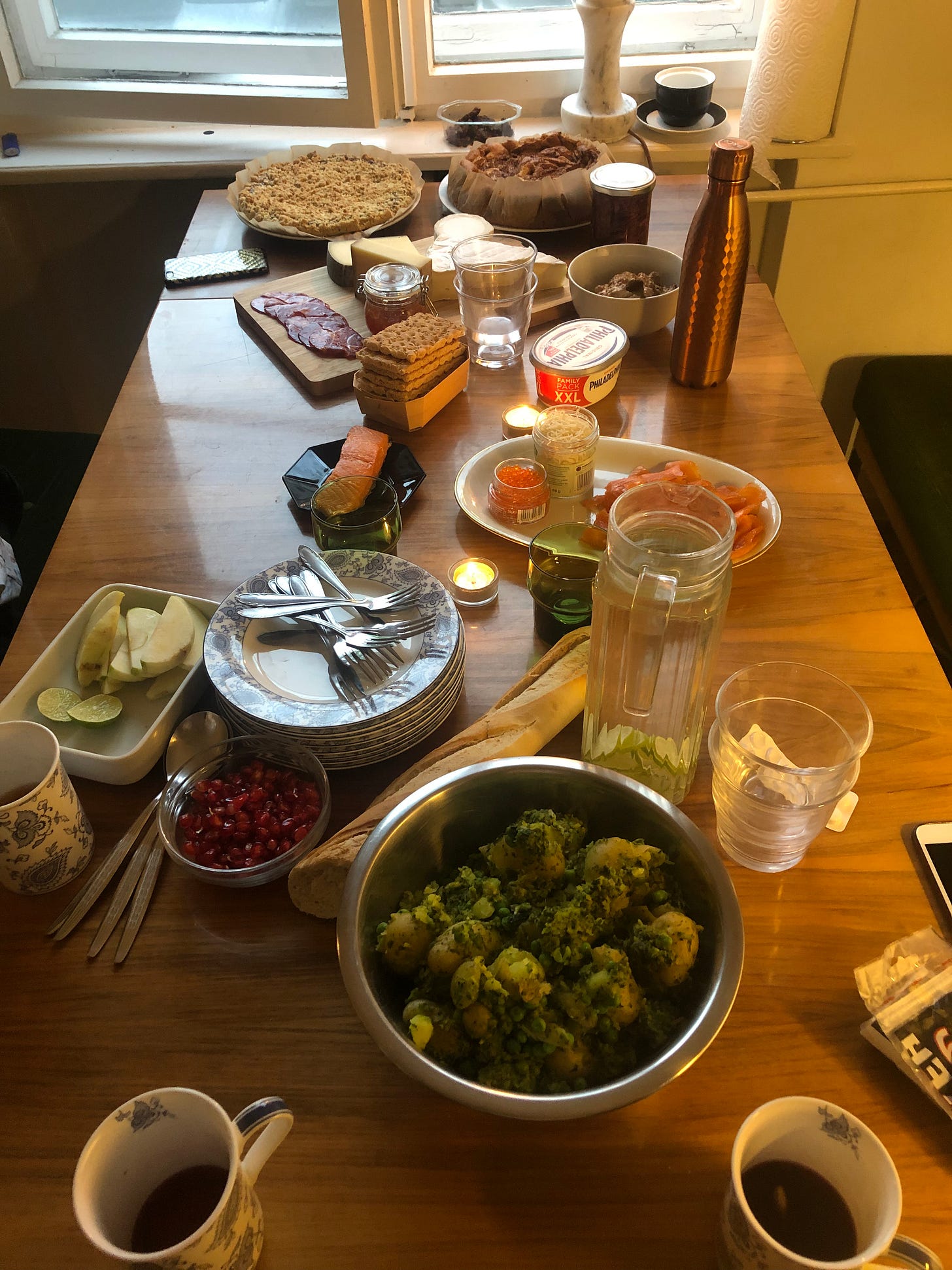 A photo from the end of a long wooden table, covered in a spread of foodstuffs, including tea cups, a large bowl of potatoes, a stack of plates and forks, a baguette, more cutlery, crackers, a cheese and meat board, and some tealights. 