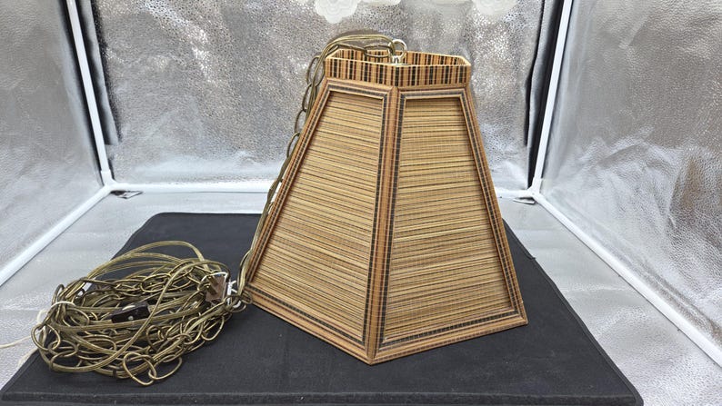 May include: A trapezoidal lampshade made of woven bamboo strips, with a dark brown frame. A brass-colored chain and electrical cord are attached. The lampshade is on a black surface, against a silver backdrop. May include: A trapezoidal lampshade made of woven bamboo strips, with a dark brown frame. A brass-colored chain and electrical cord are attached. The lampshade is on a black surface, against a silver backdrop.