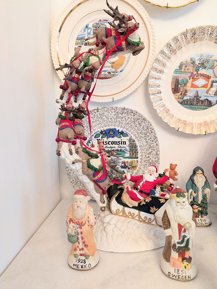 Festive display of Santa in a sleigh with reindeer, surrounded by vintage Santa figurines from Mexico, Sweden, and Yugoslavia, set against decorative plates. Festive display of Santa in a sleigh with reindeer, surrounded by vintage Santa figurines from Mexico, Sweden, and Yugoslavia, set against decorative plates.