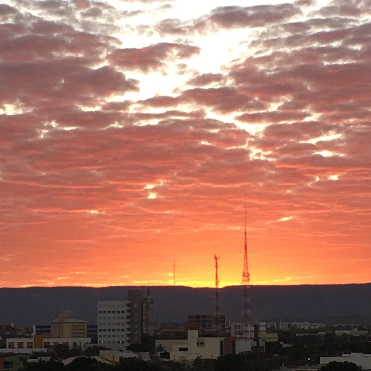 Foto do nascer do sol em dia nublado. No horizonte há uma serra, e na frente da serra, a cidade. Há duas grandes anteas. No céu, as cores formam um gradiente de um vermelho luminoso no horizonte até um vermelho cinzento no topo, tudo refletido pelas nuvens esparsas. Por trás das nuvens, se vê o céu claro.