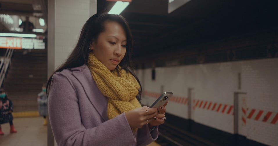  "Woman wearing a purple coat and yellow scarf stands on a subway platform, looking at her phone with a focused expression. The subway station features tiled walls with red and white safety markings, and a staircase leading to the exit is visible in the background.