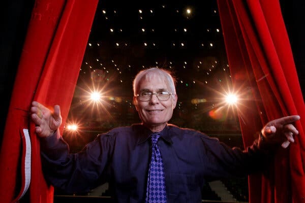 Wearing a dark shirt and tie and glasses, Speight Jenkins smiles and poses by seeming to part a red curtain and emerge out of it.