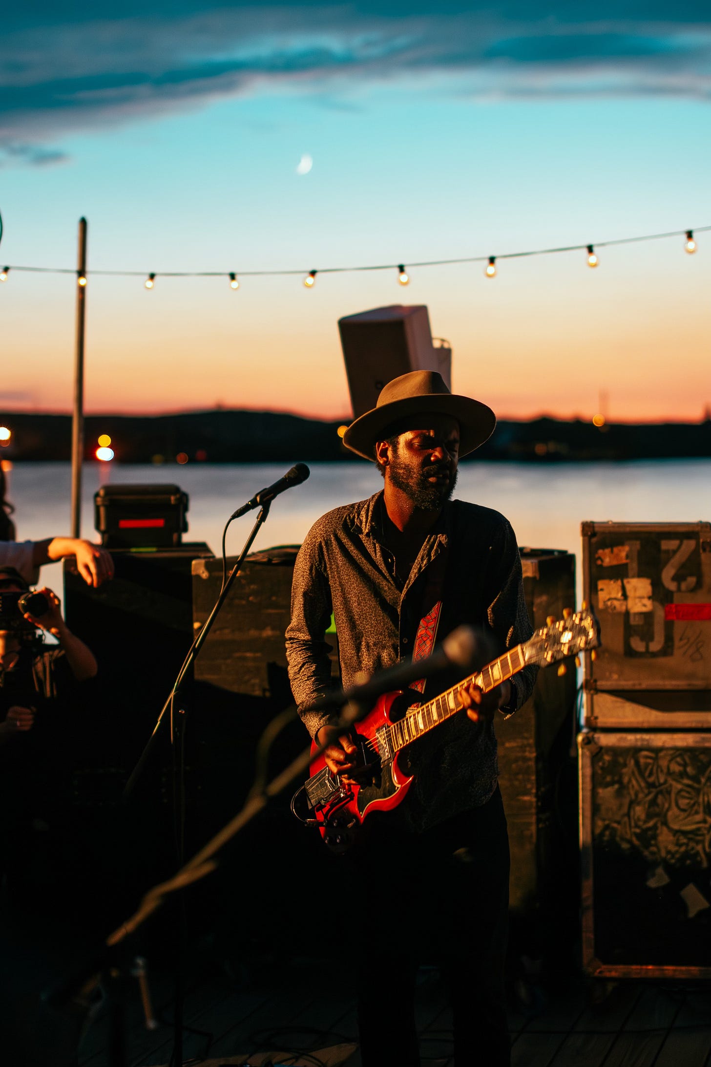Gary Clark Jr. plays his red electric guitar on an outdoor stage at dusk. He is wearing a hat and is framed by strings of lights against a twilight sky with a crescent moon visible.