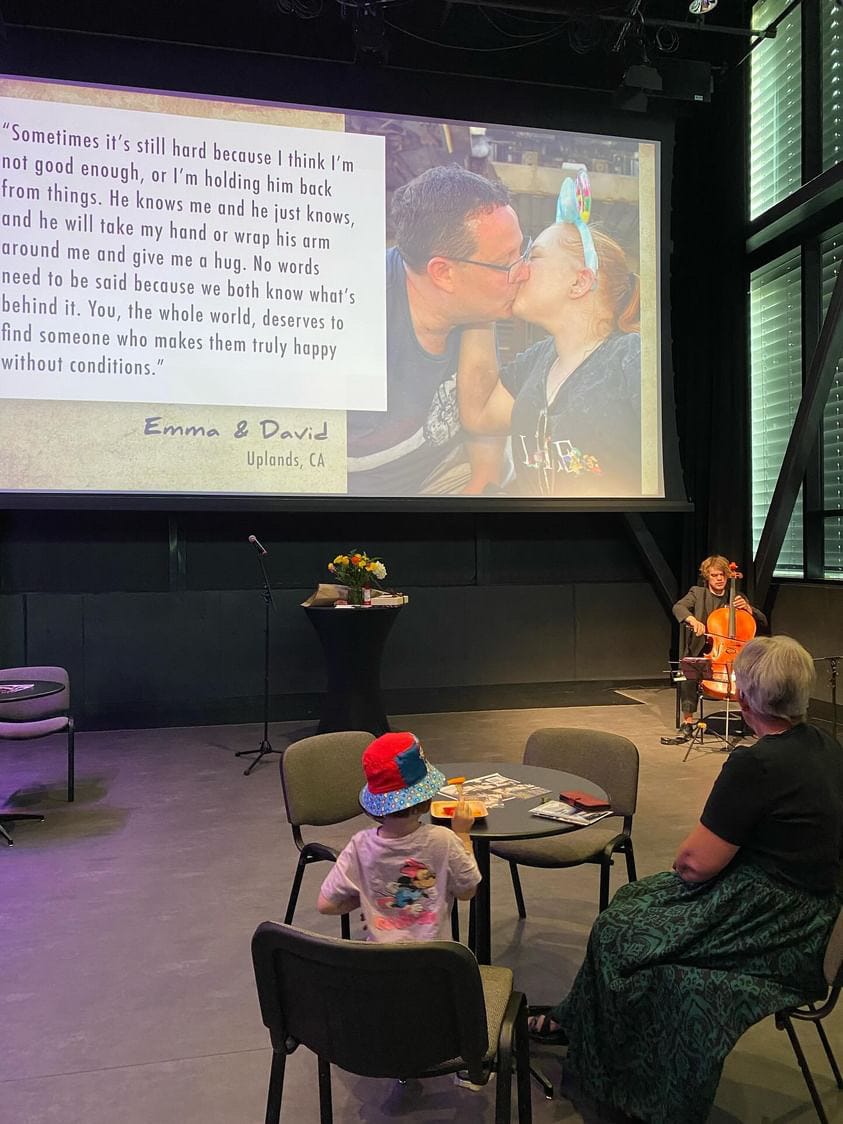 A cellist performs in front of a large projection screen with an image of a couple kissing and their story written up beside them. A cellist performs in front of a large projection screen with an image of a couple kissing and their story written up beside them.