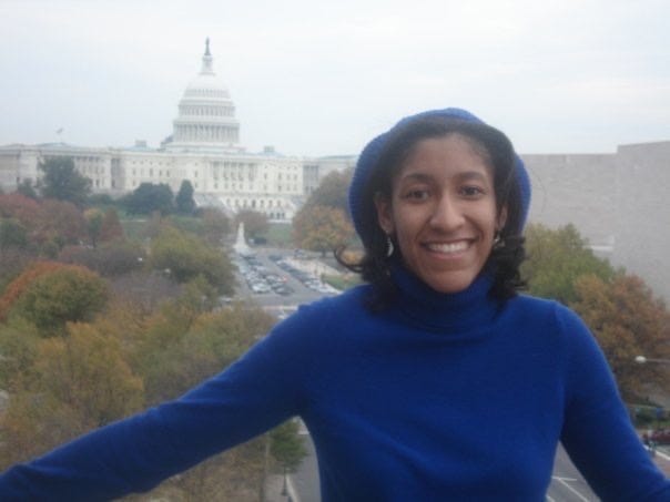 Kristen standing in a blue turtleneck and a blue sweater, with straightened but feathered Black hair on a balcony with the West front of the US Capitol building and a cloudy sky in the background. Kristen standing in a blue turtleneck and a blue sweater, with straightened but feathered Black hair on a balcony with the West front of the US Capitol building and a cloudy sky in the background.